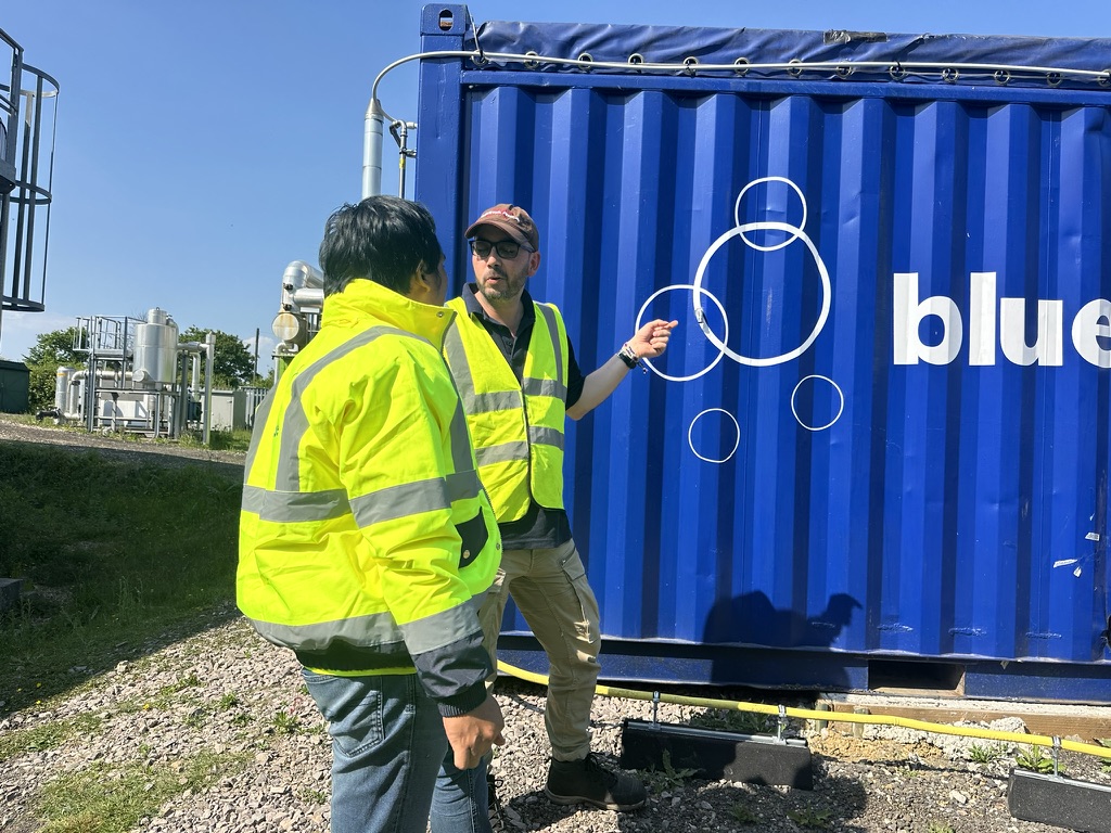 Two workers standing front of a shipping container with the bluemethane logo.