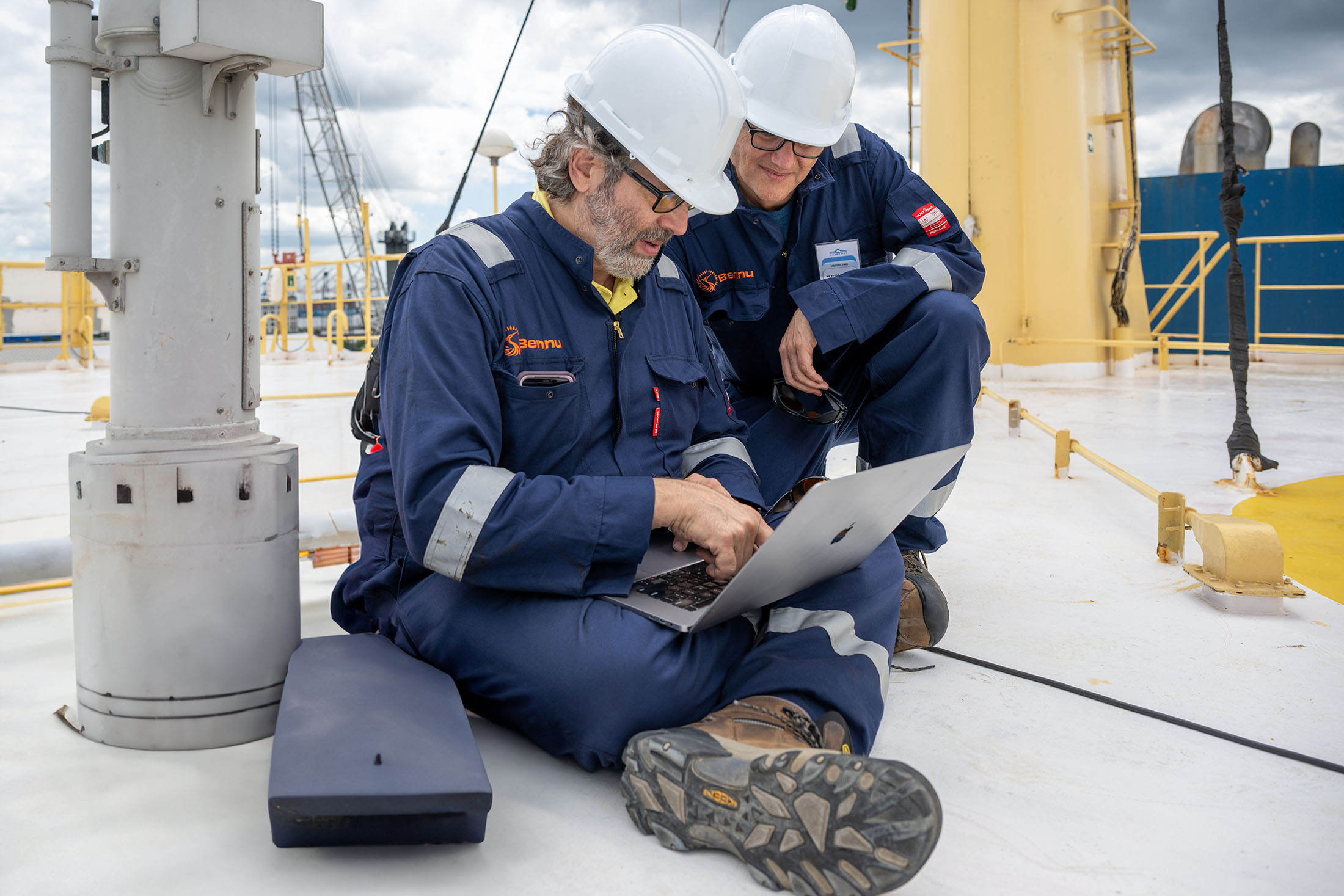 Two men on the deck of a ship looking at laptop computer. 